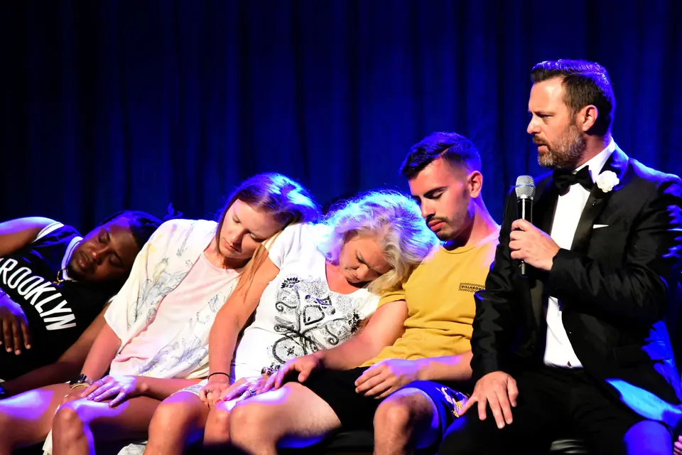 Mentalist and hypnotist Steve Falcon with a microphone beside hypnotized audience volunteers asleep on stage at Wonders Theater